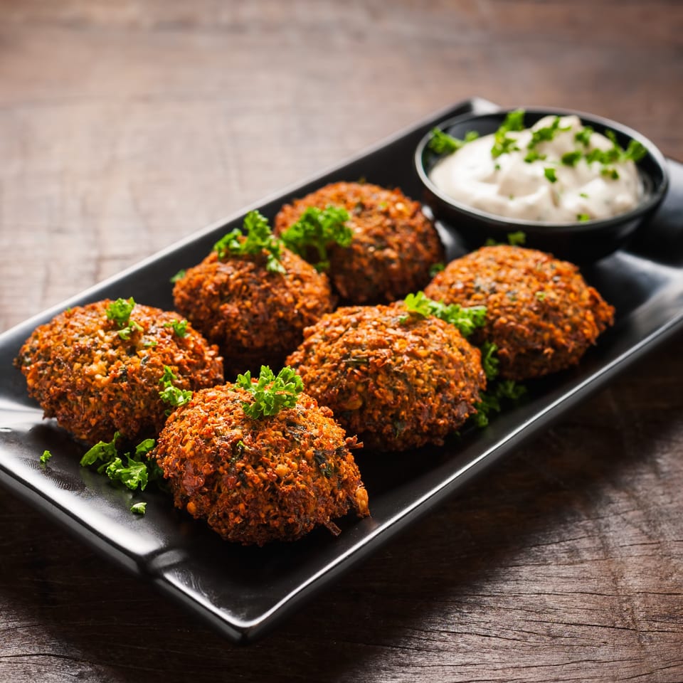 Six golden-brown falafel balls garnished with parsley, served with a side of white sauce on a black plate.