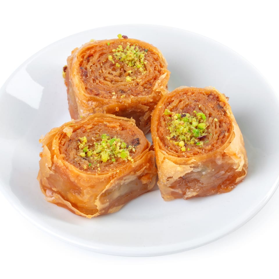 Three golden-brown middle-eastern baklava rolls, topped with green pistachios, on a white plate.